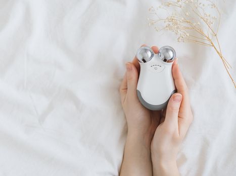 Microcurrent concept. Face massager with micro currents for home use and skincare on white bed. Female hands holds device for anti-aging and face lifting. Top view or flat lay, copy space for text.