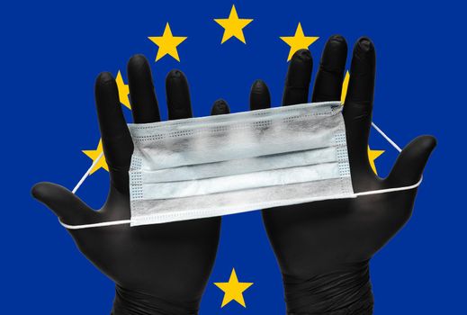 Doctor holds face mask in hands in black medical gloves on background colors flag of European Union EU. Pandemic insurance coronavirus, grippe, airborne diseases, flu. Human respiratory medical mask.