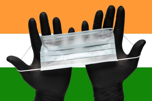 Doctor holds face mask in hands in black medical gloves on background colors National Flag of India. Pandemic insurance coronavirus, flu, airborne diseases. Human medical respiratory mask.