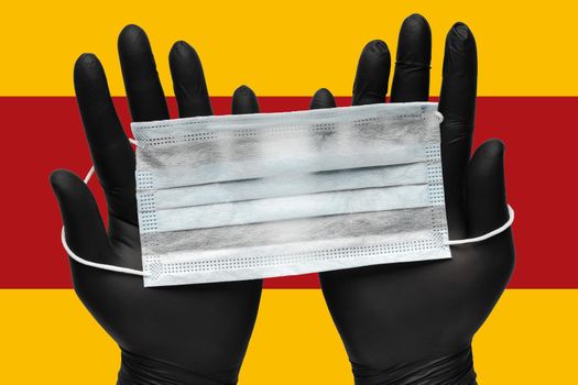 Doctor holds antivirus face mask in hands in black medical gloves on background colors flag of Spain. Pandemic insurance coronavirus, airborne diseases, SARS and grippe. Human medical respiratory mask