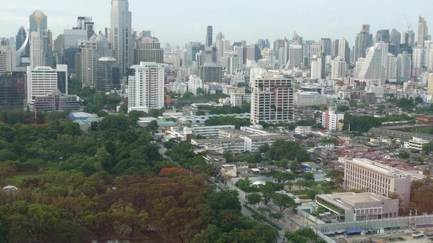 Green park in metropolitan city. Drone view of green trees of Lumphini Park near road and high rise buildings on streets of Bangkok, Thailand.