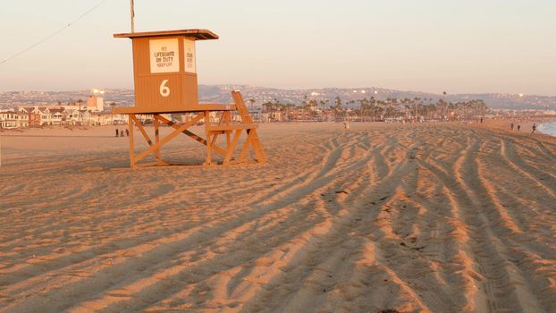 Iconic retro wooden orange lifeguard watch tower on sandy california pacific ocean beach illuminated by sunset rays. Private holiday houses and mountains on the horizon. Newport resort aesthetic, USA
