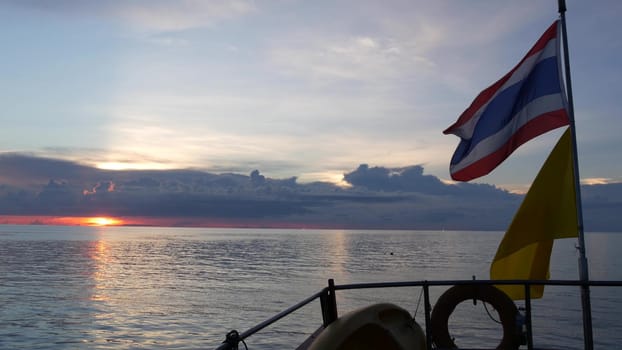 Boat floating near Thai flags in evening. Silhouette of anonymous person floating on boat on rippling sea near Thai and King of Thailand flags against cloudy sundown sky