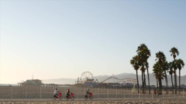 California beach aesthetic, people ride cycles on a bicycle path. Blurred, defocused background. Amusement park on pier and palms in Santa Monica american pacific ocean resort, Los Angeles CA USA.