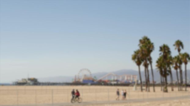 California beach aesthetic, people ride cycles on a bicycle path. Blurred, defocused background. Amusement park on pier and palms in Santa Monica american pacific ocean resort, Los Angeles CA USA.