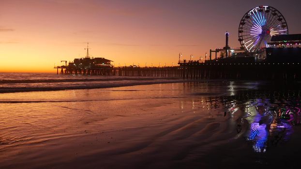 Twilight waves against classic illuminated ferris wheel, amusement park on pier in Santa Monica pacific ocean beach resort. Summertime iconic symbol of California glowing in dusk, Los Angeles, CA USA
