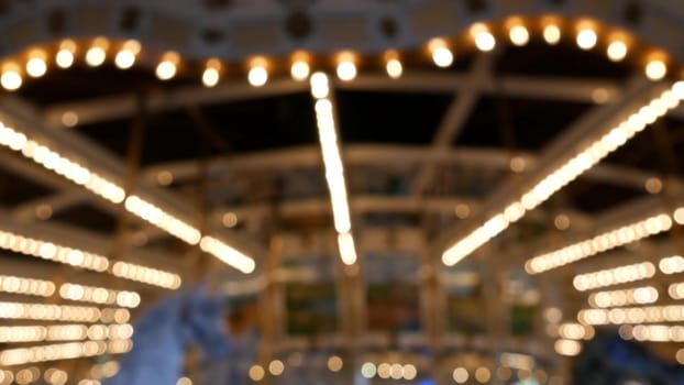 Golden illuminated defocused retro carousel turning in amusement park. Blurred shiny merry-go-round glowing and rotating at night. Radiant spinning symbol of childhood. Festival atmosphere of funfair.
