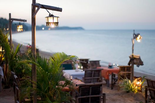 ,Resort restaurant on tropical beach in twilight, View of glowing lamps and served tables of outside restaurant on shoreline of tropical ocean, Thailand, Koh Samui.
