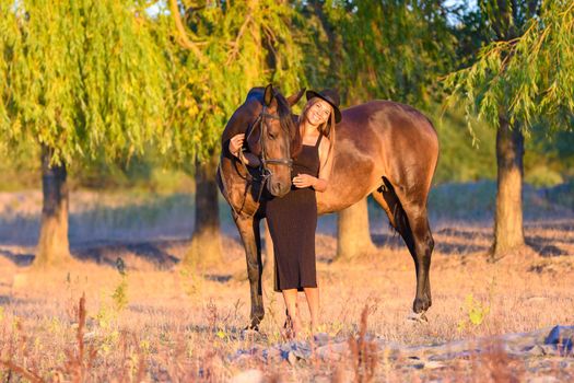 A girl with a horse stands against the background of trees, the rays of the setting sun fall on them