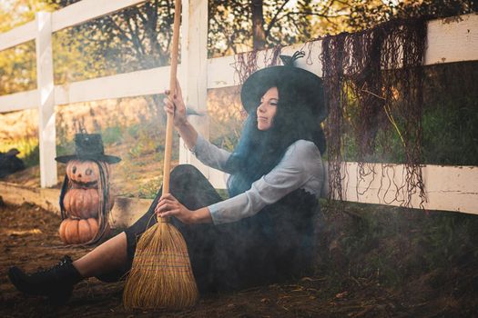 The witch sits by the fence on the farm and blows the smoke with a broom
