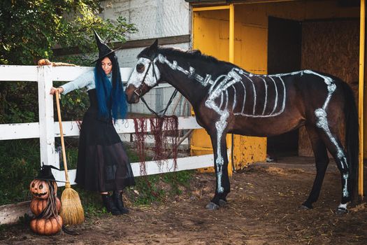 A girl dressed as a witch stands by the fence of the corral, and a horse with a painted skeleton stands next to it