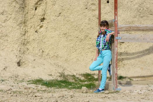 Stylish cute girl in a plaid shirt and jeans leans on a wooden structure against a background of a rock