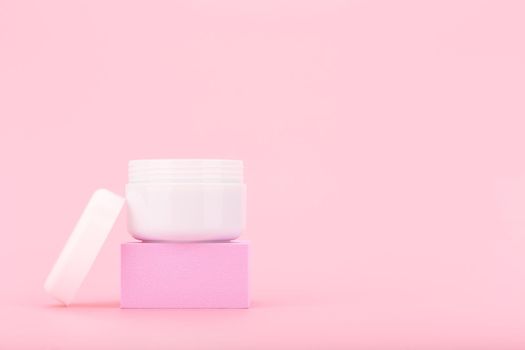 White opened cosmetic jar with cream, scrub or mask for face or hair on pink pedestal against pink background with copy space. Beauty products concept