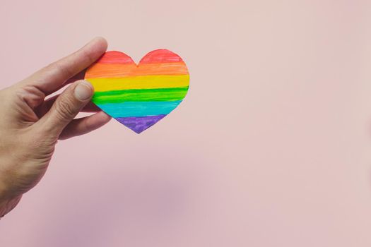 Female hands holding heart with rainbow stripes on pink background. LGBT pride flag, symbol of lesbian, gay, bisexual, transgender, social movements. Homosexual love, Human rights concept. Copy space.