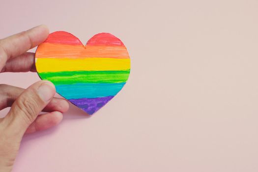 Female hands holding heart with rainbow stripes on pink background. LGBT pride flag, symbol of lesbian, gay, bisexual, transgender, social movements. Homosexual love, Human rights concept. Copy space.