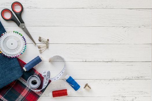 Sewing accessories, denim and plaid fabric on a white wooden background. Fabric, sewing threads, needle, pins, scissors, buttons and sewing centimeter. Copyspace