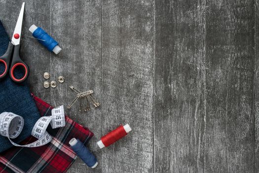 Sewing accessories, denim and plaid fabric on a dark wooden background. Fabric, sewing threads, needle, pins, scissors, buttons and sewing centimeter. Copyspace