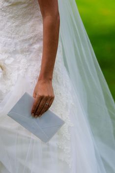 Hand of the bride holding an white enveloppe from a guest and wedding ring at her hand. Her white weddingdress is background.