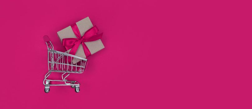 Supermarket trolley and gift box on a pink background with copy space. Shopping concept.