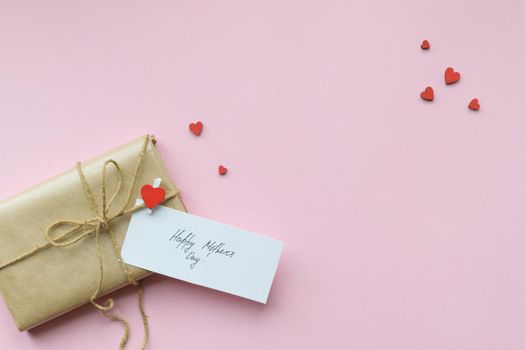 Present wrapped in brown craft paper and tie hemp string on Light pink background. Gift box with greetings on Mothers Day and decorative red hearts. Top view. Copyspace