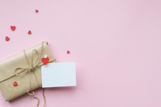 Present wrapped in brown craft paper and tie on Light pink background. Gift with decorative red hearts and empty greeting card for Mothers Day, Wedding, Birthday and Valentines Day. Top view. Copyspace. Mockup