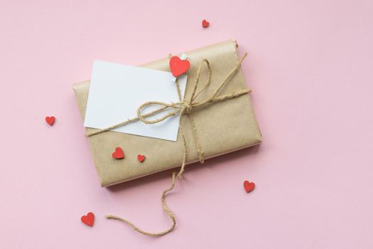 Present wrapped in brown craft paper and tie on Light pink background. Gift with decorative red hearts and empty greeting card for Mothers Day, Wedding, Birthday and Valentines Day. Top view. Copyspace. Mockup