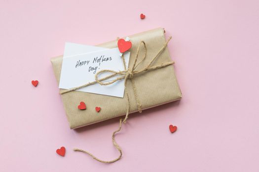 Present wrapped in brown craft paper and tie hemp string on Light pink background. Gift box with greetings on Mothers Day and decorative red hearts. Top view. Copyspace