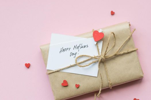 Present wrapped in brown craft paper and tie hemp string on Light pink background. Gift box with greetings on Mothers Day and decorative red hearts. Top view. Copyspace