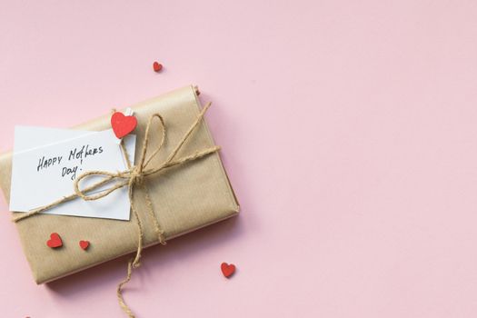Present wrapped in brown craft paper and tie hemp string on Light pink background. Gift box with greetings on Mothers Day and decorative red hearts. Top view. Copyspace