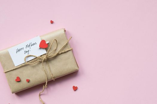 Presents wrapped in brown craft paper and tie hemp string on Light pink background. Gift box with greetings on Mothers Day and decorative red hearts. Top view. Copyspace