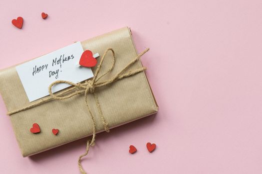 Present wrapped in brown craft paper and tie hemp string on Light pink background. Gift box with greetings on Mothers Day and decorative red hearts. Top view. Copyspace