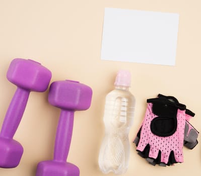 pink sports gloves, pair of purple dumbbells and and bottle of water on a beige background, top view, healthy lifestyle