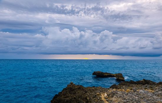 A calm sunset on a dark blue sea. Thick clouds on the horizon. In the foreground is a rocky shore. Romantic mood.