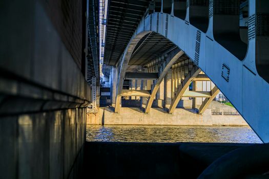 Powerful iron bridge structure, View from below. Concrete pillars, big iron rivets. A river flows under the bridge. Perspective.