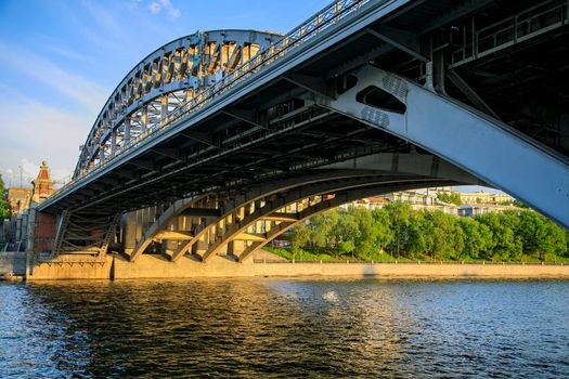 Powerful iron bridge structure, View from below. Concrete pillars, big iron rivets. A river flows under the bridge. Perspective.