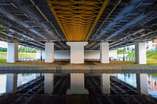 Powerful iron bridge structure, View from below. Concrete pillars, big iron rivets. A river flows under the bridge. Perspective.