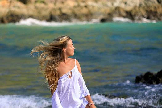 A young beautiful girl looks at the sea, the wind develops her blond hair. White dress, blue water., beautiful view.