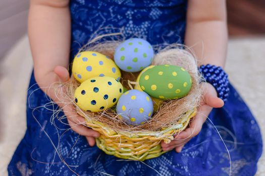 A basket with colorful Easter eggs in the hands of a little girl in a blue dress. Celebration of the Holy Easter. 