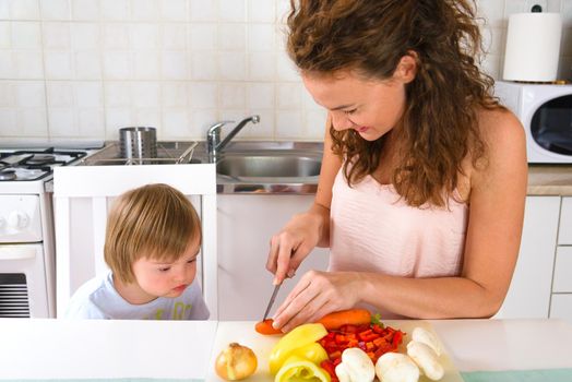 Young women preparing healthy launch from vegetables in white kitchen while his son toddler with Down Syndrome curiously looking. In white kitchen.