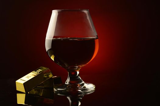 A concept of wealth utilizing a couple gold bullion bars next to a rich Cognac drink over a dark reflective red and black background.