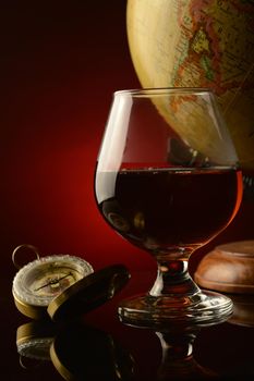A shallow depth of field focus on a glass of Cognac with a compass and antique globe with a dark reflective red and black background.