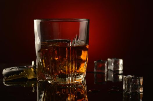 A concept image of drinking and driving utilizing a set of car keys next to a glass of whiskey over a dark reflective black and red background.