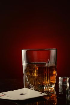 A vertical composition of a whiskey drink and a made poker hand over a dark reflective red and black background.