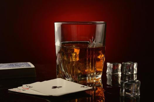 A glass of whiskey next to a deck of playing cards and a made poker hand revealed over a dark reflective red and black background.