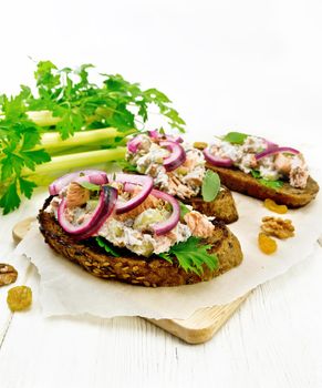 Salmon, petiole celery, raisins, walnuts, red onions and curd cheese salad on toasted bread with green lettuce leaves on parchment on a wooden board background