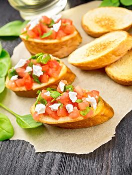 Bruschetta with tomato, spinach and soft cheese on parchment, garlic, vegetable oil in a decanter and basil on dark wooden board background