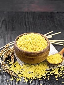 Bulgur groats - steamed wheat grains - in a clay bowl and a spoon on sacking, ears of wheat on the background of wooden board