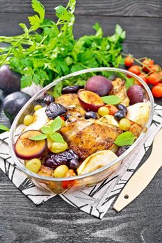 Baked chicken with tomatoes, apples, plums and grapes in a glass roaster on a kitchen towel, garlic, parsley and basil on dark wooden board background