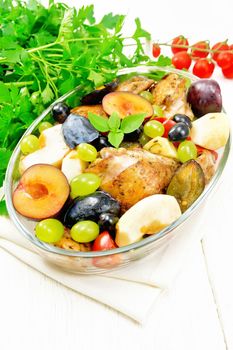 Baked chicken with tomatoes, apples, plums and grapes in a glass roaster on a light napkin, garlic, parsley and basil on white wooden board background