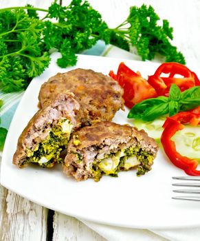 Cutlets stuffed with spinach and egg, salad with tomatoes, cucumber and pepper in a dish on a napkin, basil and parsley on the background light wooden boards